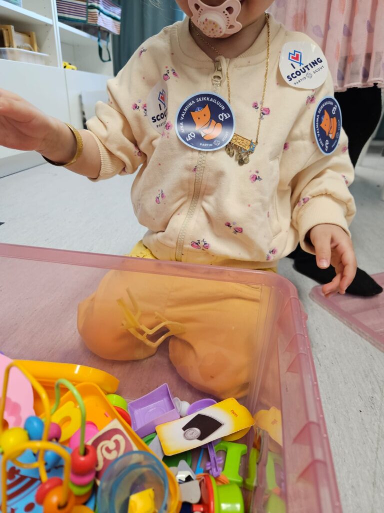 Kuvassa lapsi polvi-istunnassa. Paitaan kiinnitetty partio-tarroja, joissa lukee "aina valmiina seikkailuun".